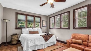 Ein Schlafzimmer mit einem Bett, einem Deckenventilator, einem Fenster mit Jalousien, einem Nachttisch und einem Ledersessel.