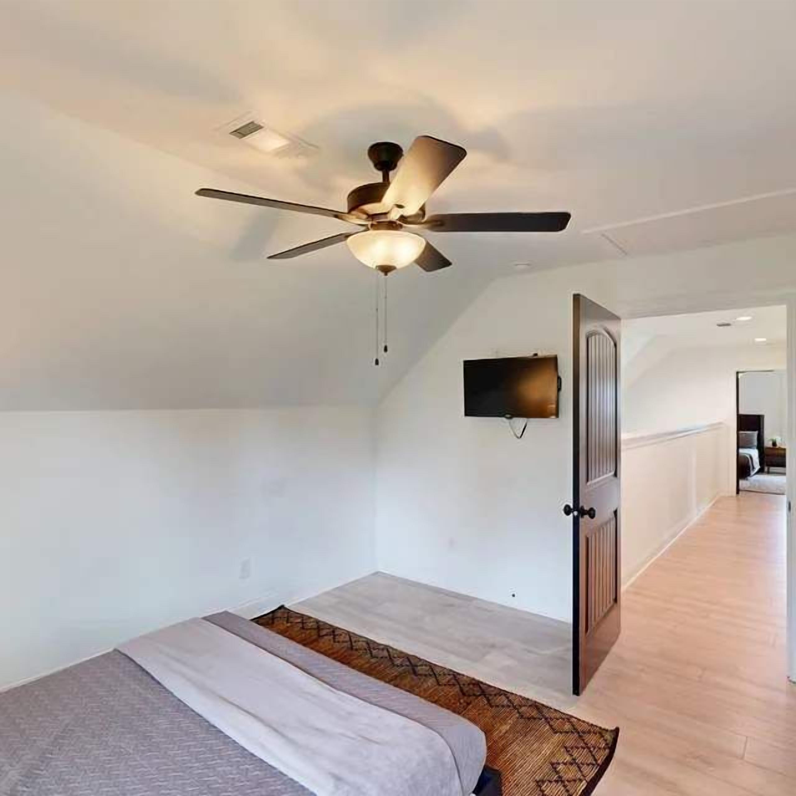 A room with a bed, a ceiling fan, a TV mounted on the wall, and a door leading to another room.