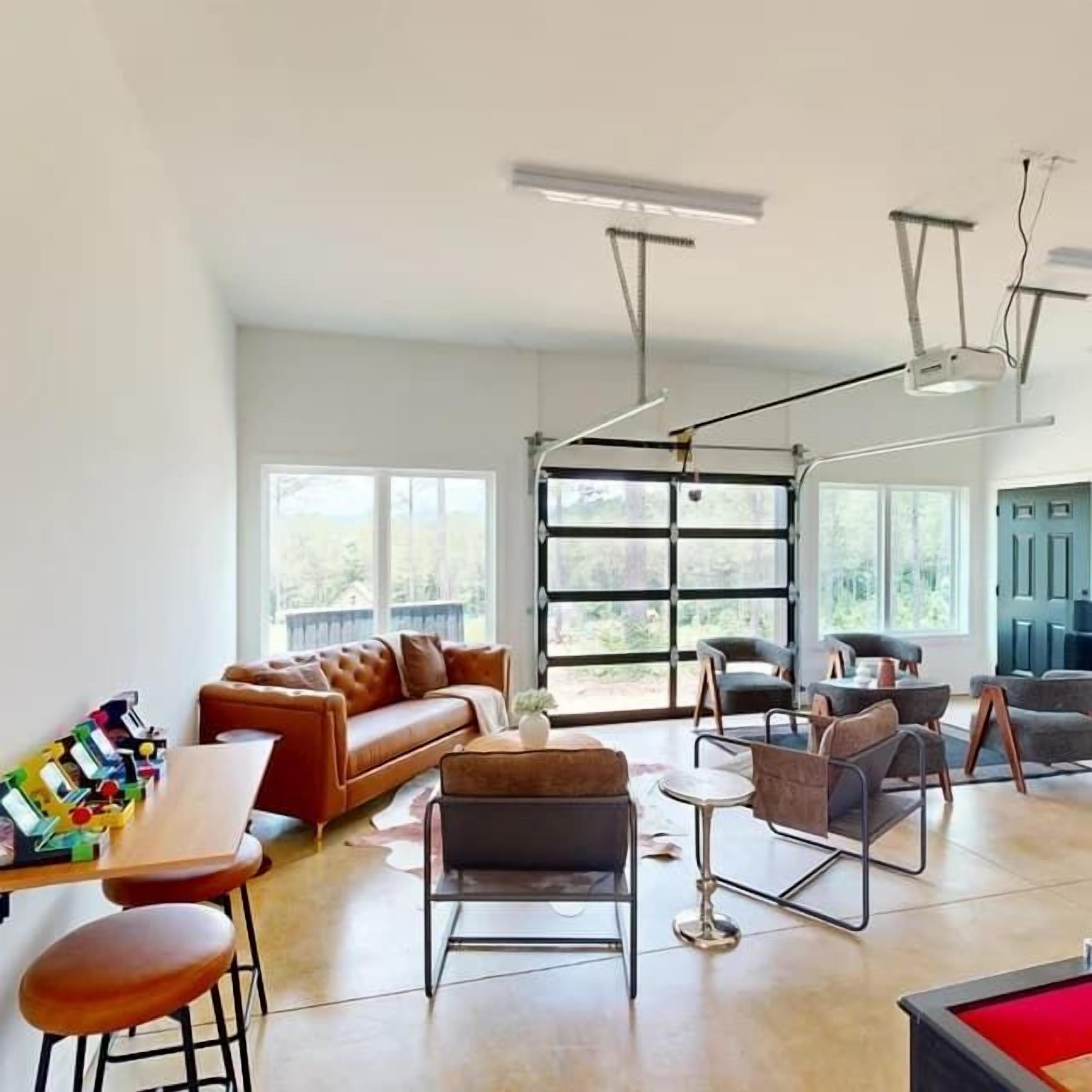 A spacious living area with a brown leather sofa, a coffee table, and modern chairs. There is a large window providing natural light and a view of the outdoors.