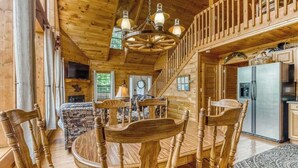 Interior de una cabaña de madera con una mesa de comedor, sillas, una araña, una zona de cocina y una escalera.