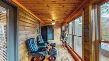 A log cabin interior with a wooden ceiling, a ceiling fan, a seating area with leather chairs and ottomans, and large windows offering a view of the outdoors.