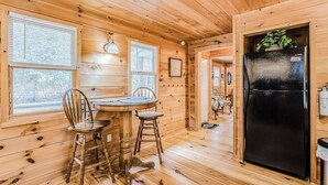 Ein Interieur einer Holzhütte mit einem Esstisch, Stühlen, einem schwarzen Kühlschrank und einem Fenster mit Jalousien.