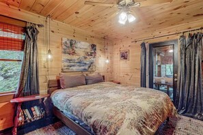 A wooden cabin bedroom with a bed, a nightstand, a window with curtains, a ceiling fan, and a painting on the wall. - It's a Wonderful Life (Sautee Nacoochee)