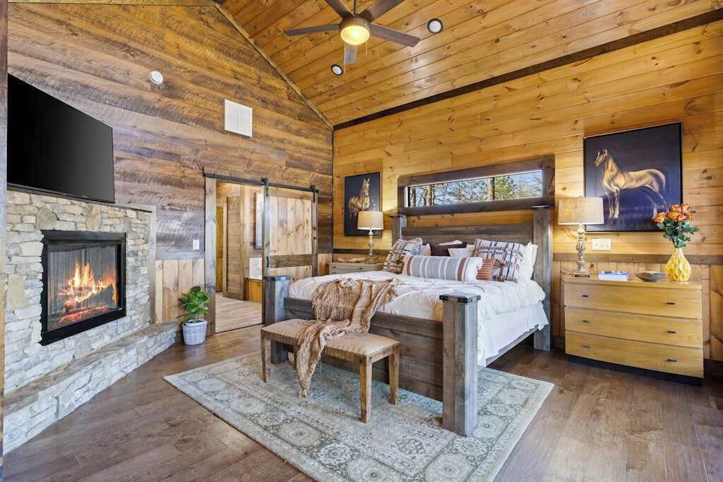 Un dormitorio con chimenea de piedra, techo de madera, una cama con almohadas, un banco y una mesita de noche.