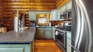 A log cabin interior with a kitchen featuring granite countertops, a stainless steel refrigerator, and a wood-burning stove. - Lakeview Manor Cabin (Blue Ridge)