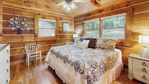 Camera da letto in una baita di legno, con un letto, una sedia, un comodino, una ventola a soffitto e finestre.