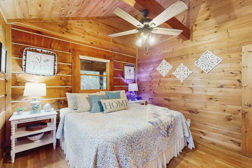 Een slaapkamer in een houten hut met een bed, nachtkastjes, een plafondventilator en decoratieve wandversieringen.