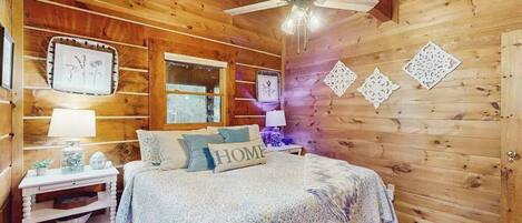Een slaapkamer in een houten hut met een bed, nachtkastjes, een plafondventilator en decoratieve wandversieringen.