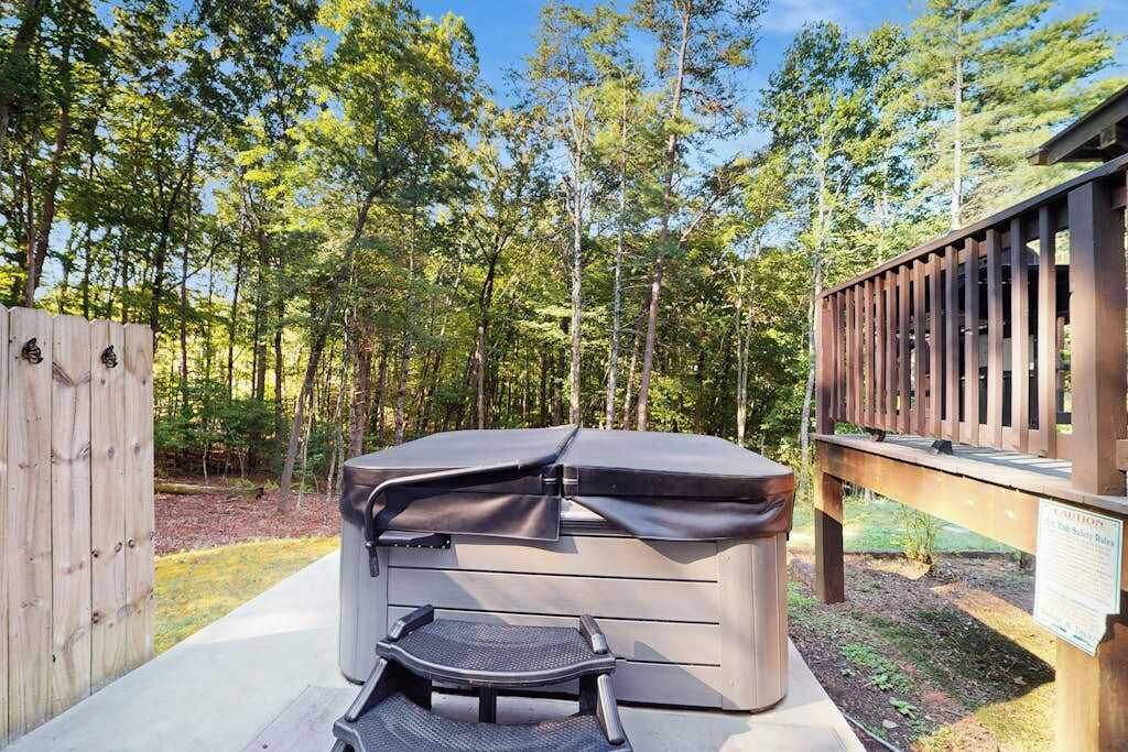 A hot tub with a cover, a chair, and a wooden deck in a wooded area.