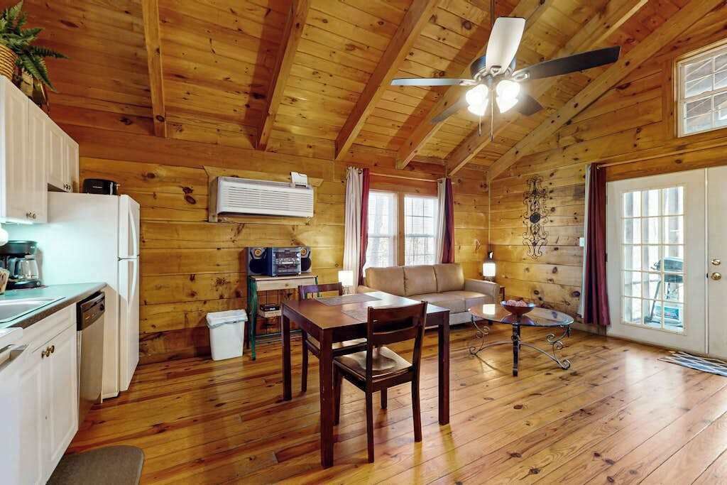 A wooden cabin interior with a dining table, chairs, a sofa, a ceiling fan, and a kitchen area.