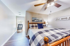 A bedroom with a bed, a chair, a ceiling fan, a painting, and a wall sign.