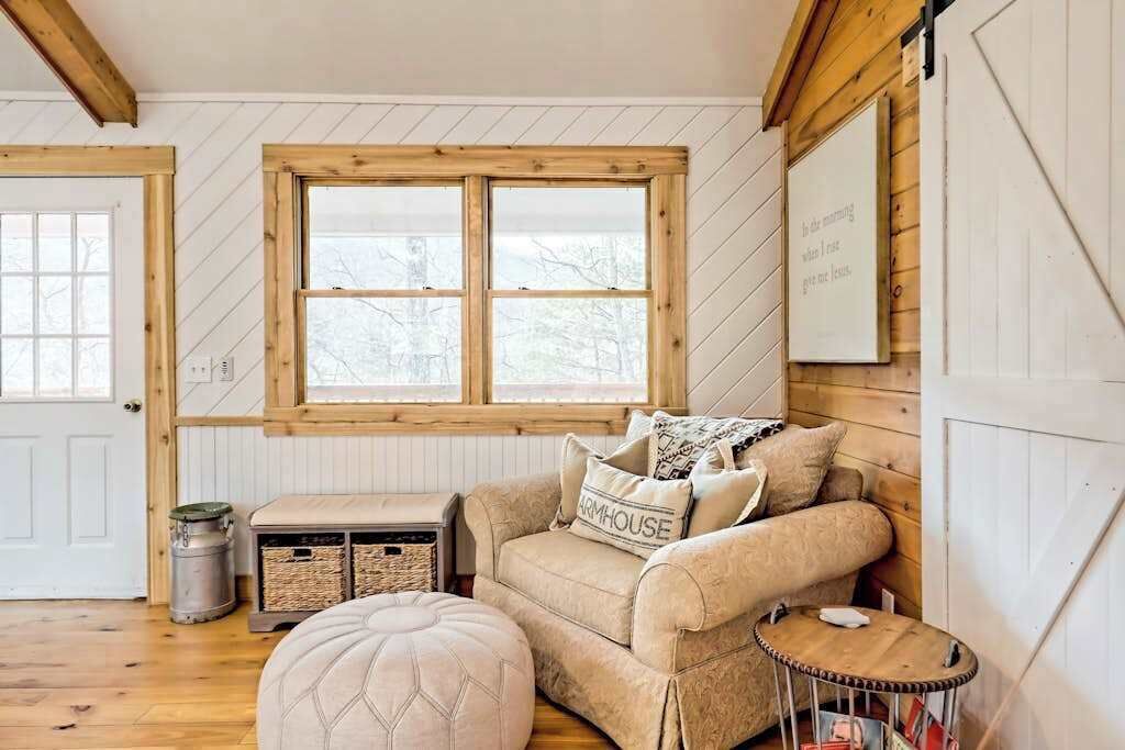 Ein gemütliches Zimmer mit einem raumhohen Holzfenster, einer gepolsterten Couch, einem kleinen Hocker und einem an der Wand befestigten Schild.