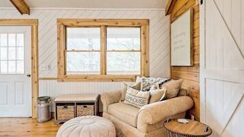 Ein gemütliches Zimmer mit einem raumhohen Holzfenster, einer gepolsterten Couch, einem kleinen Hocker und einem an der Wand befestigten Schild.