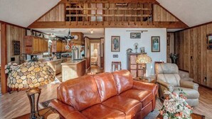 A spacious loft-style living area with a high ceiling, wooden beams, a leather sofa, a dining table, and a kitchen in the background. - The Bird House Stay - Firepit and Grill (Dillard)