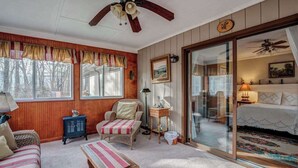 A room with a bed, a chair, a small table, a window with curtains, a ceiling fan, and a sliding glass door leading to another room. - The Bird House Stay - Firepit and Grill (Dillard)