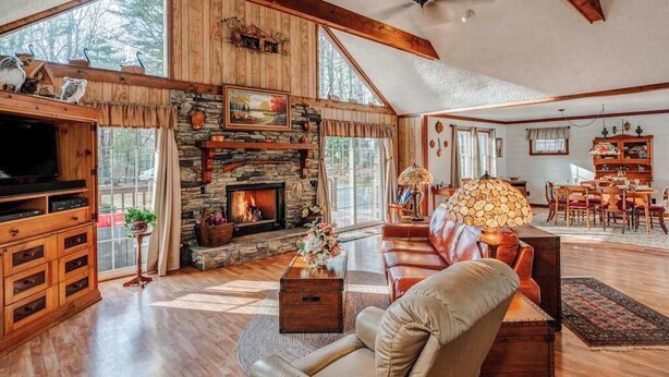A spacious living room with a stone fireplace, wooden beams, a television, and a cozy seating area. - The Bird House Stay - Firepit and Grill (Dillard)