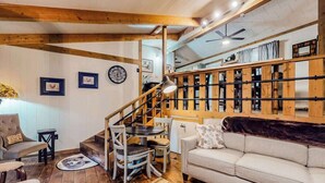 A cozy living room with a staircase, wooden beams, a sofa, a dining table, and a ceiling fan. - White Pine Perch Stay (Clayton)