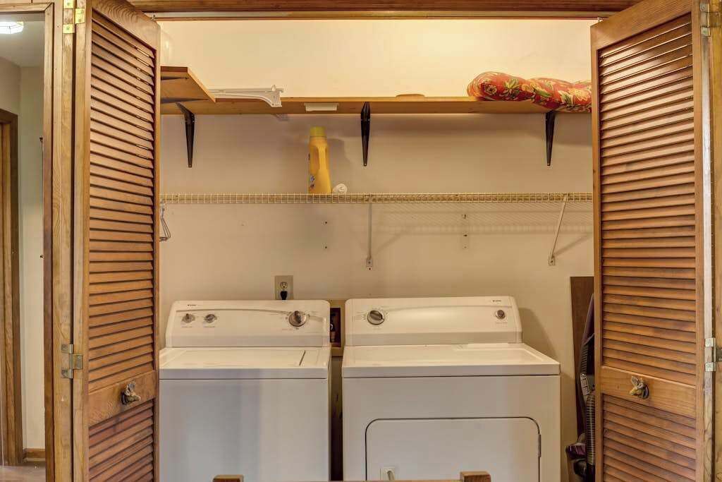 A laundry room with two white washing machines, wooden slatted walls, and a shelf with a yellow bottle and a black object.