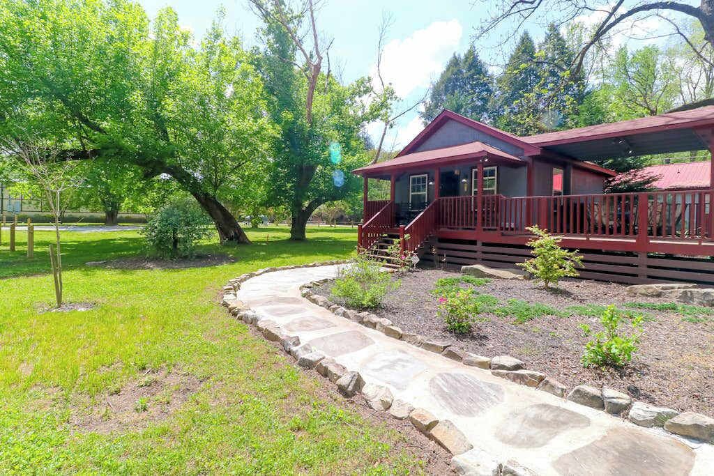 A wooden cabin with a stone pathway leading to the entrance, surrounded by lush greenery and trees.