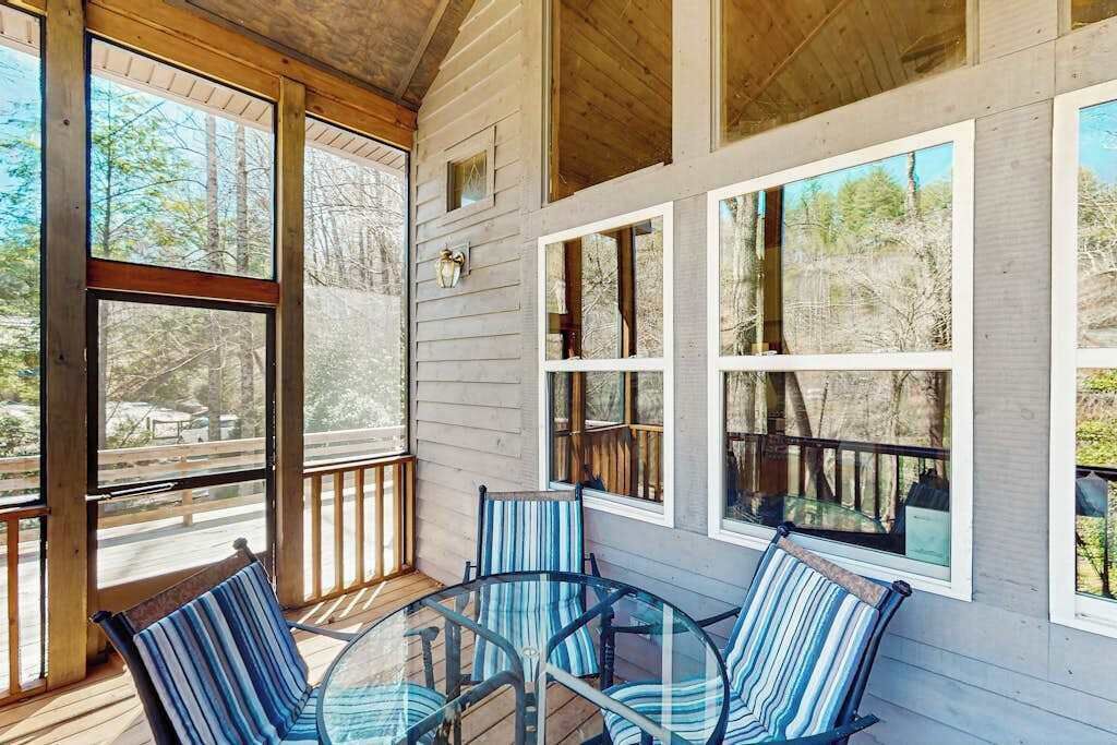Une véranda avec une table en verre et des chaises à rayures, des balustrades en bois et de grandes fenêtres donnant sur les arbres.