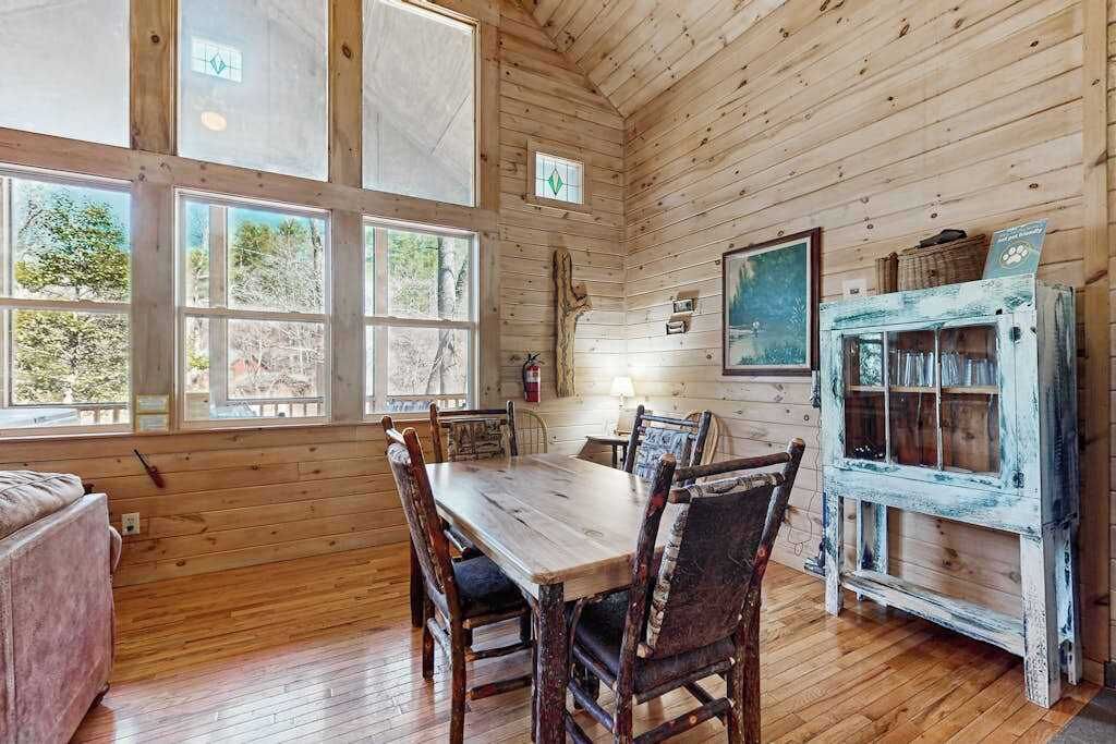 A wooden cabin interior with a dining table, chairs, a small fireplace, and a rustic cabinet.