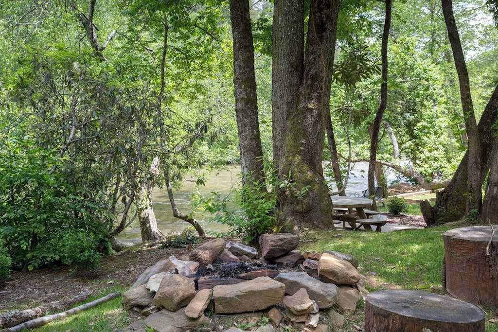 A forested campsite with a fire pit, picnic table, and a view of a lake.