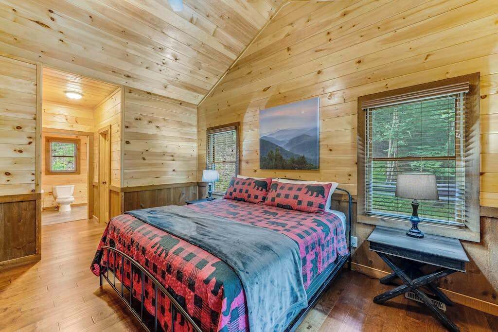 Un dormitorio de cabaña de madera con una cama, una mesita de noche, una ventana con persianas y un cuadro en la pared.