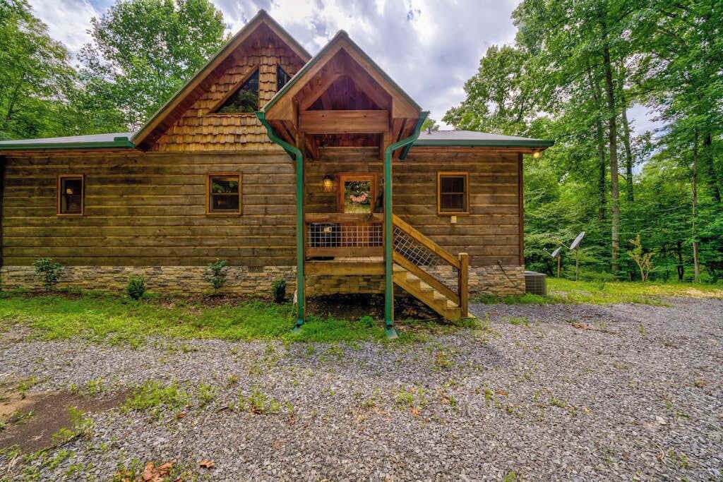 A rustic log cabin with a wooden staircase, a front porch, and a gravel driveway.