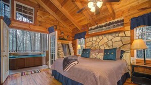 A log cabin bedroom with a stone fireplace, a large bed, a bench by the window, and a ceiling fan.