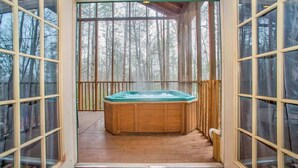 A wooden hot tub on a deck with glass doors, surrounded by a forest.