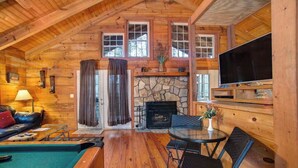A log cabin interior with a stone fireplace, a television, a pool, and a dining area.