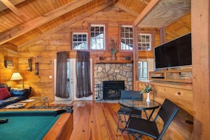 Interior de una cabaña de troncos con chimenea de piedra, televisor, piscina y comedor.
