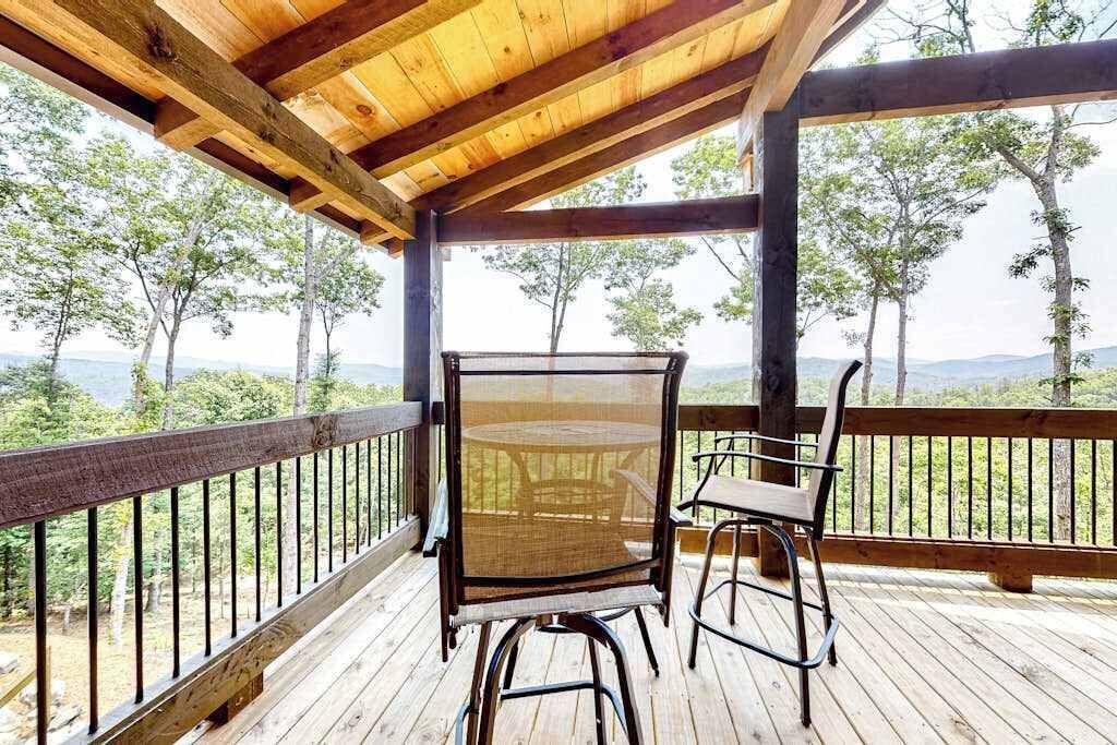 A wooden deck with a table and chairs, overlooking a forested area with trees and a distant view of mountains.