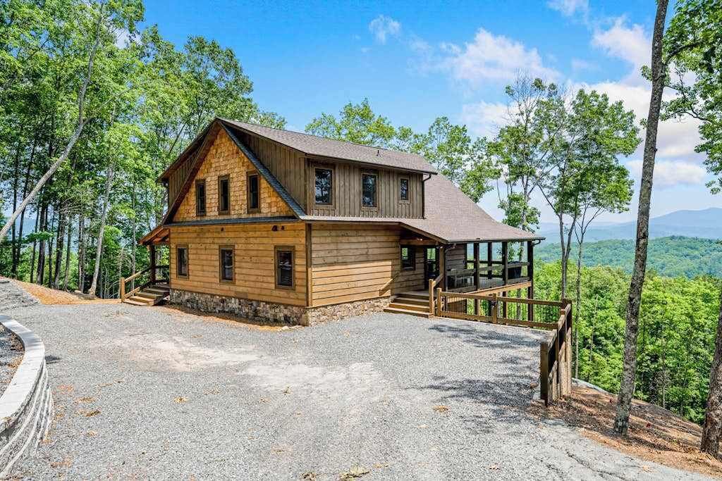 A wooden cabin with a porch, surrounded by trees and a gravel driveway.
