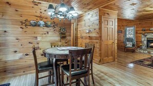 Ein Interieur einer Holzhütte mit Esstisch, Stühlen, einer Deckenleuchte, einem Kamin und einer Tür zu einem weiteren Raum.