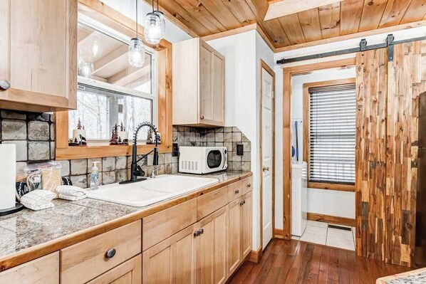 A kitchen with wooden cabinets, a stone backsplash, a microwave, and a hanging light fixture. - Wooded Wonder Retreat (Cleveland)