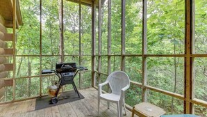 A covered outdoor area with a grill, a white chair, and a view of the forest.