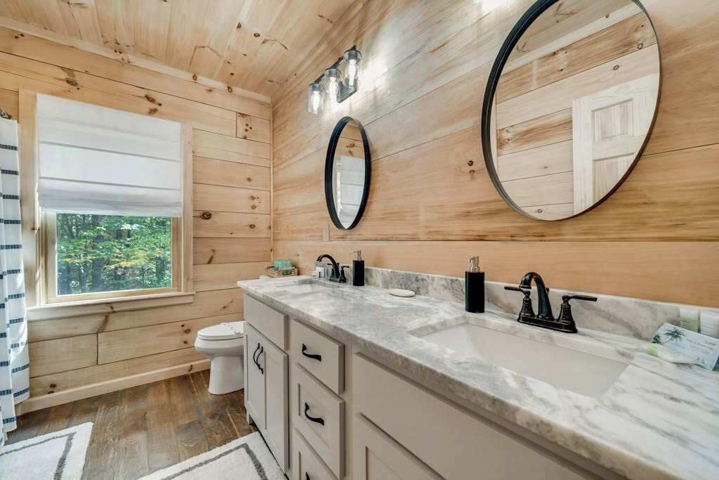 Baño de cabaña de madera con un ventanal grande, un lavabo con encimera de mármol y dos espejos redondos.