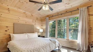 Un dormitorio de cabaña de madera con una cama, un ventilador de techo, una mesita de noche y una ventana con cortinas.