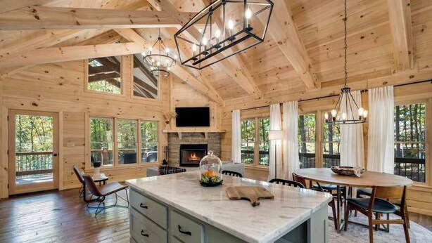 Un interior de cabaña espacioso con una amplia mesa de madera para comer, una cocina con chimenea de piedra y un ventanal con vistas al bosque.