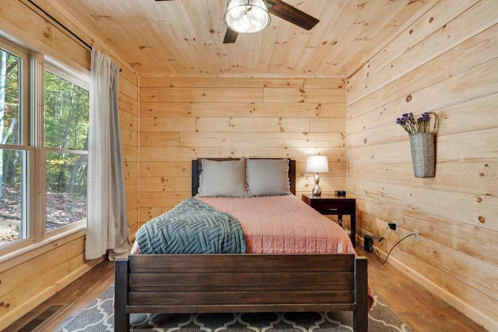 A wooden-paneled bedroom with a bed, nightstand, lamp, and window.