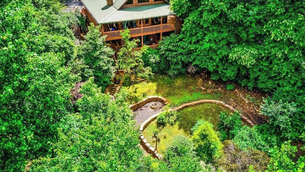Une grande maison en bois, entourée d’une végétation luxuriante, avec un étang visible et un sentier en pierre.
