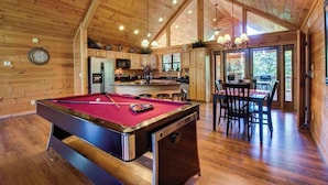 Un intérieur en bois comprenant une table de billard, une cuisine, un coin repas et de grandes fenêtres.