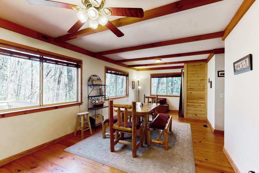 A room with a wooden ceiling, large windows, a dining table with chairs, and a ceiling fan.
