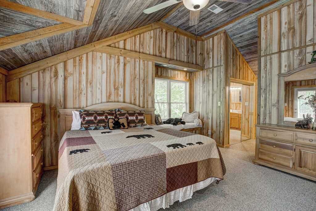 A bedroom with a wooden ceiling, a bed with a quilt, a dresser, and a window.