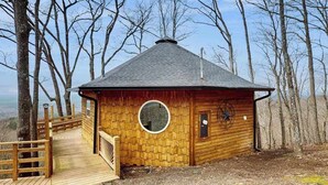 Une cabane en bois dotée d’une fenêtre ronde, d’une terrasse en bois et d’une porte en métal.