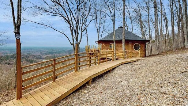 Une cabane en bois dotée d’une fenêtre circulaire, entourée d’arbres, et un chemin de bois menant à cette cabane.