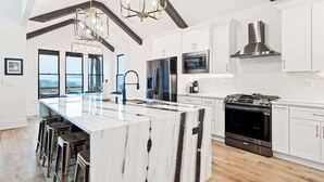 A modern kitchen with a large island, stainless steel appliances, and a high ceiling with exposed beams. - Knight Mountain Stay (Ellijay)