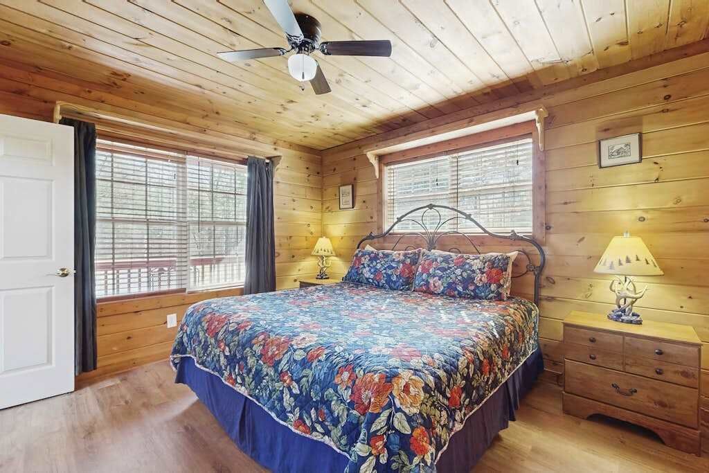 Un dormitorio de cabaña de madera con una cama, dos ventanas, un ventilador de techo y una mesita de noche.