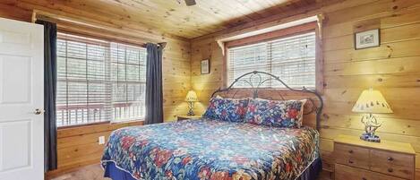 Un dormitorio de cabaña de madera con una cama, dos ventanas, un ventilador de techo y una mesita de noche.